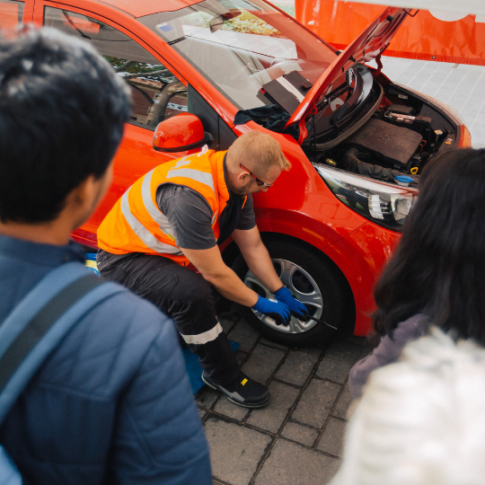 A man in an orange hi-vis vest demonstrates how to change a tyre on a red car