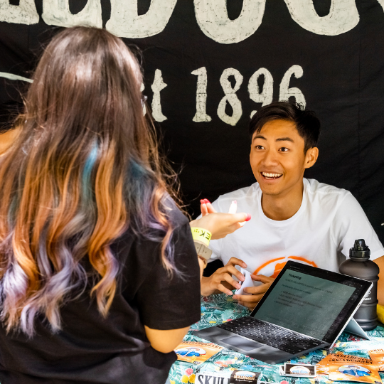 A student with long hair talks with a student in a white t-shirt seated behind an O'Week stall with a black banner